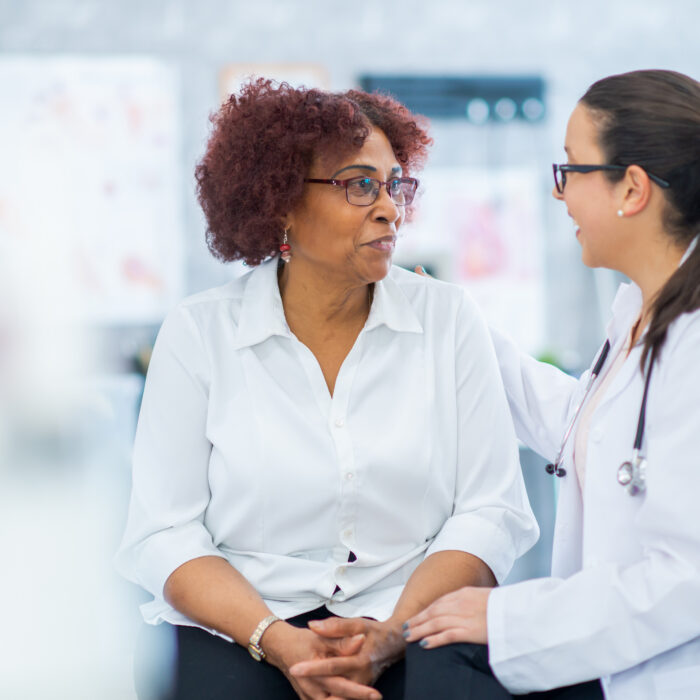 A senior woman of African decent, sits up on an exam table as she discusses her medications with her doctor. The patient is dressed casually and has a neutral expression on her face as she talks with her doctor.