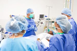 Lead surgeon is explaining surgical procedure to interns in teaching hospital while prepping patient for surgery. Doctors are wearing sterile blue surgical gowns, caps, masks, and gloves. Patient is lying on operating table.