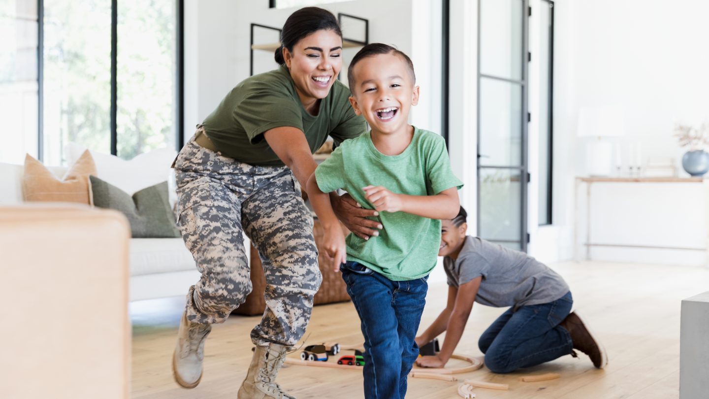While the preteen boy sets up the wooden train set on the living room floor, the female soldier chases the elementary age boy through the house. Everybody is smiling and happy.