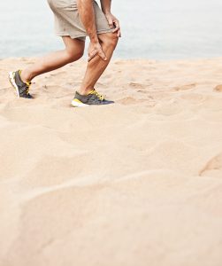 Muscle injury. Man with sprain thigh muscles. Athlete in sports shorts clutching his thigh muscles after pulling or straining them while jogging on the beach.