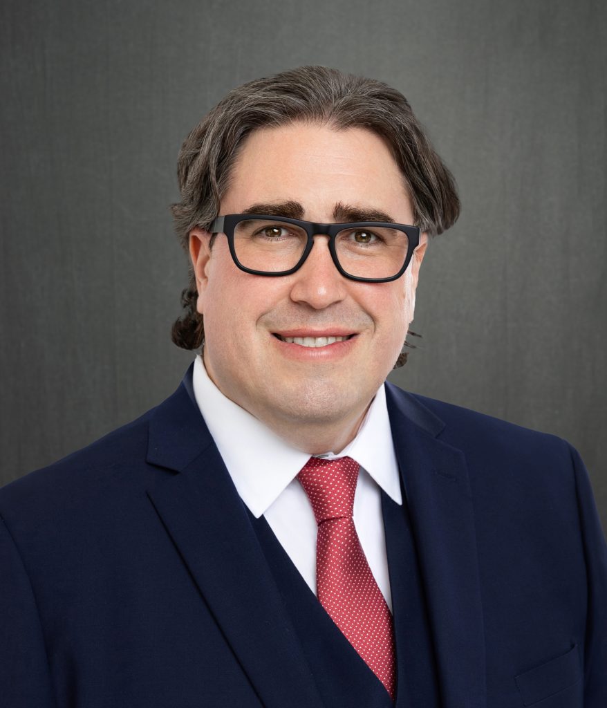 white male with glasses wearing a. navy blue suit and red tie