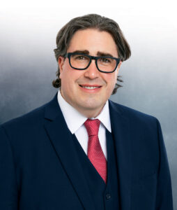 white male with glasses wearing a. navy blue suit and red tie