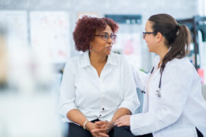 A senior woman of African decent, sits up on an exam table as she discusses her medications with her doctor. The patient is dressed casually and has a neutral expression on her face as she talks with her doctor.