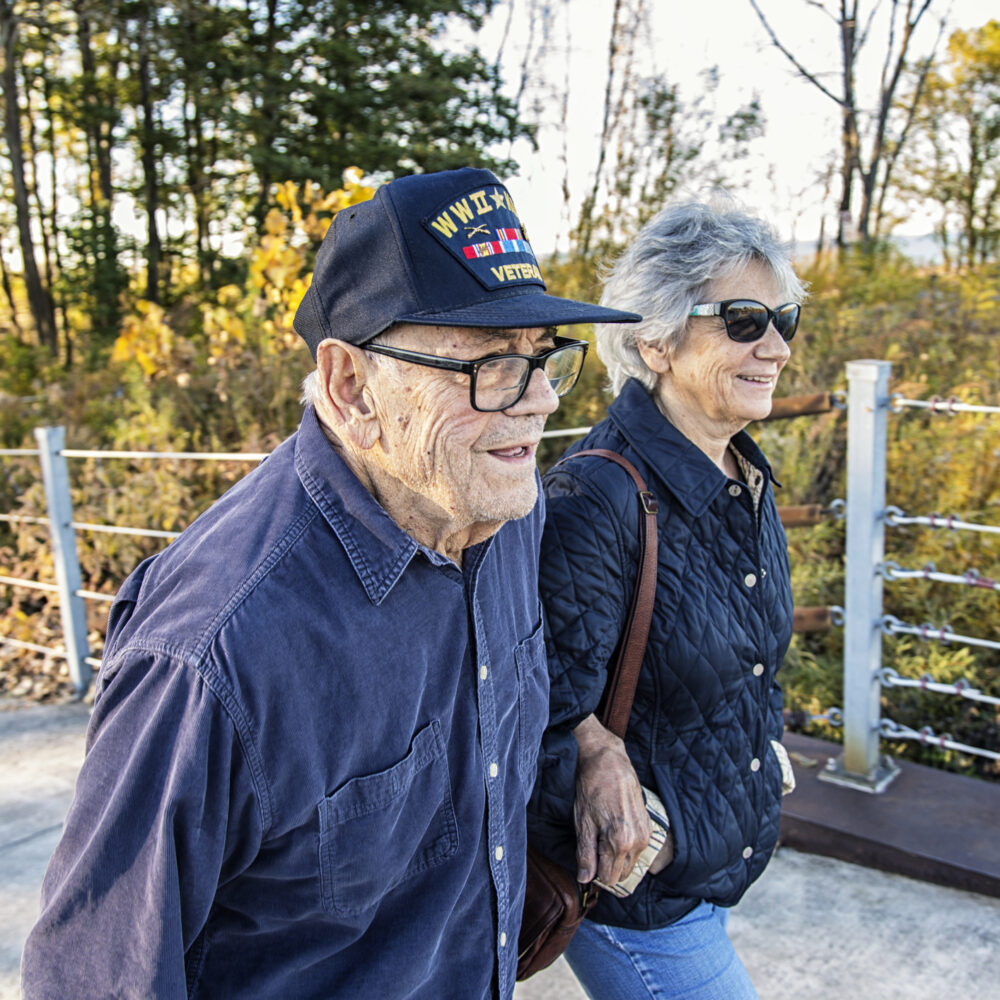 A proud, elderly, 93 year old real person senior adult man USA WWII and Korean Conflict military war veteran and his real life senior adult woman home caregiver daughter are walking side by side sightseeing on a sunny day at a public park on the shoreline of Lake Ontario. He is wearing a common, unbranded, generic souvenir shop military veteran commemorative baseball style cap with generic wording, replica plastic insignia pins and replica campaign ribbon iron-on patches. Photo taken near Rochester, N.Y., in the Finger Lakes region of western New York State, USA.<br /> NOTE: There are no official or authentic military uniform elements and no intent to portray anything other than a patriotic United States military veteran.